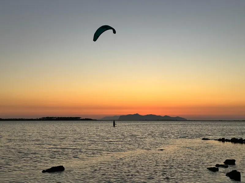 Un kite verde al tramonto sulla laguna dello Stagnone, il kitesurfista in piedi nell'acqua e le montagne all'orizzonte